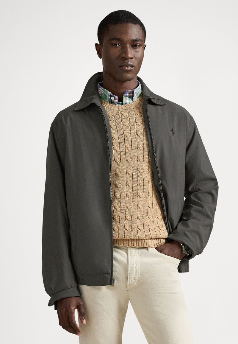 Chaqueta gris oscuro con cremallera y cuello, usada sobre un suéter de punto beige texturizado con trenzas y una camisa a cuadros, combinada con vaqueros claros.