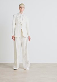 Witte krijtstrepen kostuum met een fitted blazer en wijde broekspijpen. Stof is glad met verticale strepen. Staat tegen een neutrale achtergrond.