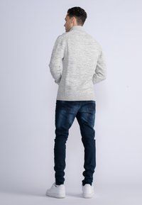 Maglione grigio chiaro a maglia con colletto alto, polsini e orlo a coste, abbinato a jeans in denim blu scuro e sneakers bianche.