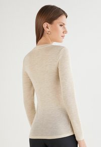 Beige Langarmshirt aus leichtem, strukturiertem Stoff mit Rundhalsausschnitt und figurbetonter Silhouette, vor einem einfarbigen Hintergrund platziert.