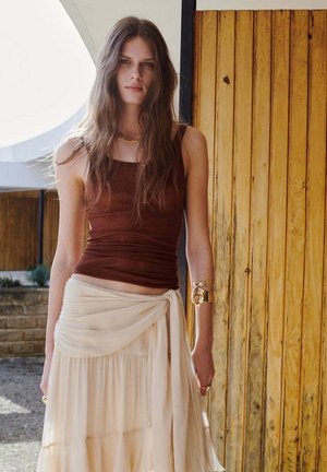 Joven mujer con largo cabello castaño lleva una blusa marrón sin mangas, falda beige de capas con detalle de envoltura, collar dorado y brazalete espiral dorado en el brazo.