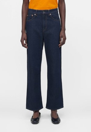 Personne portant un jean bleu foncé à jambes larges, des chaussures noires sans lacets et une chemise orange rentrée contre un fond clair uni.