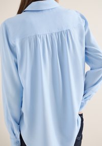 Blouse bleu clair à manches longues avec des plis froncés sur le haut du dos, portée par une personne aux cheveux foncés et en pantalon noir.