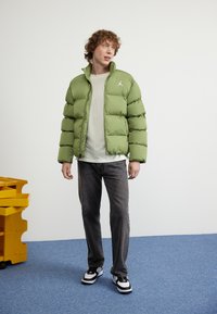 Giacca piumino verde con zip frontale e colletto alto, abbinata a una camicia leggera e jeans scuri. Presenta un accento con logo e tessuto testurizzato.