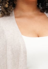 Světlý béžový kardigan z měkké látky s otevřeným předním dílem a texturovaným povrchem, vrstvený přes bílý top.