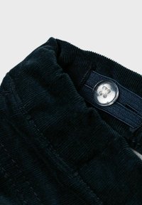 Collo in corduroy verde scuro con dettaglio di bottone argentato e etichetta in tessuto blu. Texture morbida con cuciture visibili e forma arrotondata.