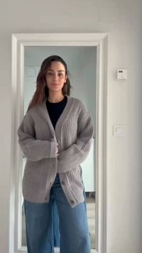 Femme debout à l'intérieur en train de boutonner un cardigan gris en tricot, portant une chemise noire et un jean bleu à jambes larges, encadrée par une porte blanche.