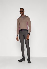 Tilpasset cykeldragt i brun med pink detaljer, lange ærmer, fuld lynlås, parret med sorte sko og solbriller. Glat tekstur, atletisk design.