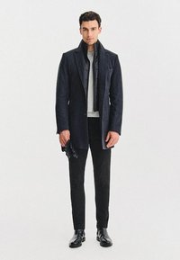 Czarny wełniany płaszcz nałożony na pikowaną kurtkę, w zestawieniu z szarym swetrem i czarnymi spodniami. Czarne rękawiczki i botki za kostkę dopełniają całość.