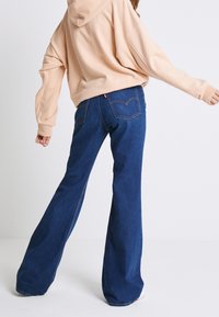 Jeans in denim blu a zampa d'elefante con lavaggio medio, caratterizzati da cuciture a contrasto, due tasche posteriori e un'etichetta rossa sulla vita.