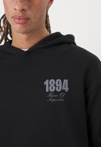 Sweatshirt à capuche noir avec un graphique sur le devant en gris clair, affichant "1894" en gras et "Maison de Superbia" en écriture cursive.