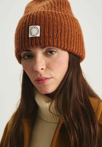 Bonnet en tricot marron avec une texture côtelée et un petit patch présentant un logo. Le modèle porte un col roulé crème et un manteau marron.