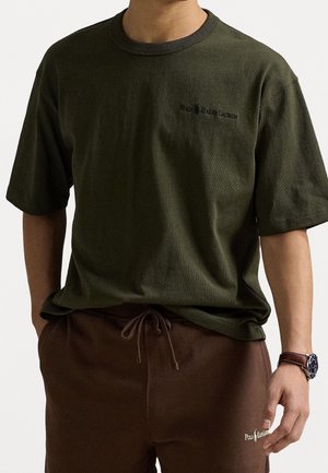 Olijfgroene katoenen T-shirt met korte mouwen, ronde hals en geborduurd logo. Draagt erbij een bruine joggingbroek met aantrekkoorden.