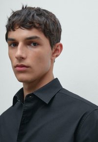 Uomo giovane con capelli corti e mossi di colore scuro, che indossa una camicia nera con bottoni, rivolto leggermente a sinistra su uno sfondo semplice e chiaro.