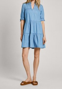 Žena v světlomodrých džínových šatech se sklady a ohrnutými rukávy, v hnědých plochých sandálech, stojící na jednom barevném pozadí.