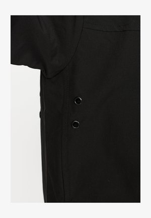 Zwarte kledingstuk met een gestructureerde stof, een ronde halslijn en cirkelvormige zwarte knopen aan de zijkant. Eenvoudige ontwerpelementen zijn duidelijk zichtbaar.