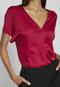 Blusa rossa in satin con scollatura a V e maniche corte, caratterizzata da una texture liscia e una vestibilità morbida. Abbinata a pantaloni neri a vita alta.