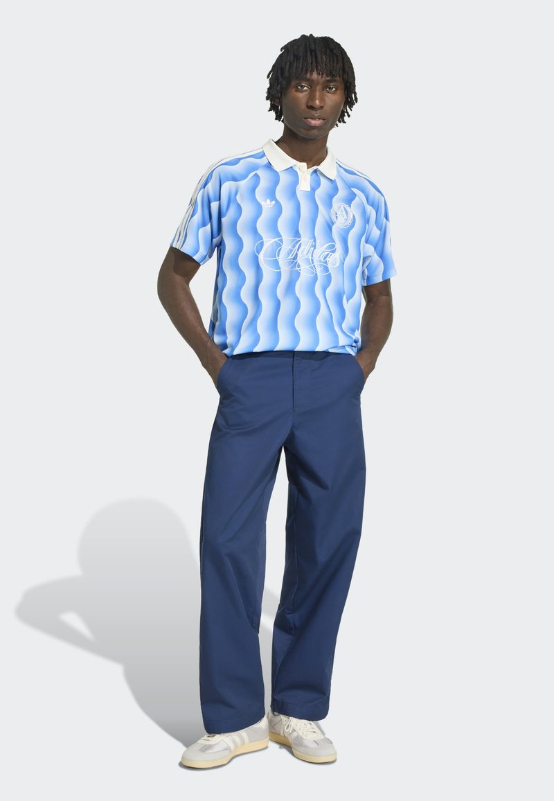 Mann steht mit Händen in den Taschen, trägt blau gemustertes Polohemd, marineblaue Hose und weiße Turnschuhe vor grauem Hintergrund.