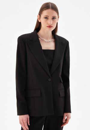 Zwarte op maat gemaakte blazer van een gladde stof, met een geknipt kraag, sluiting met een knoop aan de voorkant en twee voorzakken. Geweven zilveren kettinghalsband.