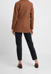 Persona con un abrigo marrón, pantalones negros y zapatos marrones de pie con la espalda hacia la cámara contra un fondo blanco liso.