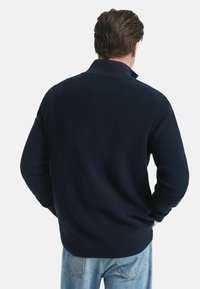 Ein Mann trägt einen dunkelblauen, gerippten Pullover mit hohem Kragen und hellblauen Jeans. Er steht mit dem Rücken zu einem einfarbigen Hintergrund.