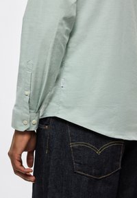 Ανοιχτό πράσινο πουκάμισο με μακριά μανίκια και μανσέτες με κουμπιά, συνδυασμένο με σκούρο τζιν με κίτρινη ραφή και κλασική σχεδίαση τσέπης.