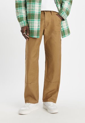 Braune Baumwollhose mit geradem Bein, vorderen Taschen und verstärkten Nähten, kombiniert mit einem grün karierten Hemd und weißen Turnschuhen.