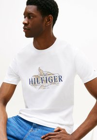 Camiseta de algodón blanca con un diseño floral en azul y dorado con el texto "HILFIGER". Manga corta y cuello redondo. Corte casual.