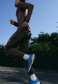 Brązowa koszula z długim rękawem z niebieskim akcentem, brązowe spodnie sportowe z kieszeniami, czerwono-niebieskie buty do biegania i białe skarpetki, na tle niebieskiego nieba.