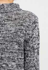 Maglione lavorato a maglia in bianco e nero, caratterizzato da un alto colletto a costine e un motivo a zigzag testurizzato sul tessuto, che mette in risalto un aspetto morbido e accogliente.
