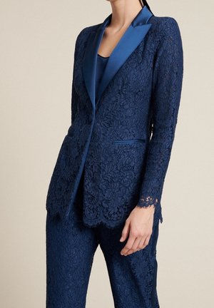 Vrouw draagt een marineblauw kanten pak met satijnen revers, bestaande uit een getailleerd blazer en bijpassende broek tegen een effen beige achtergrond.