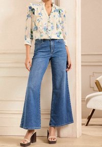 Leichte Bluse mit floralem Muster und Rüschen, geknöpft, kombiniert mit hochtaillierten, weit geschnittenen blauen Jeans. Das Model trägt beige Plateau-Sandalen.