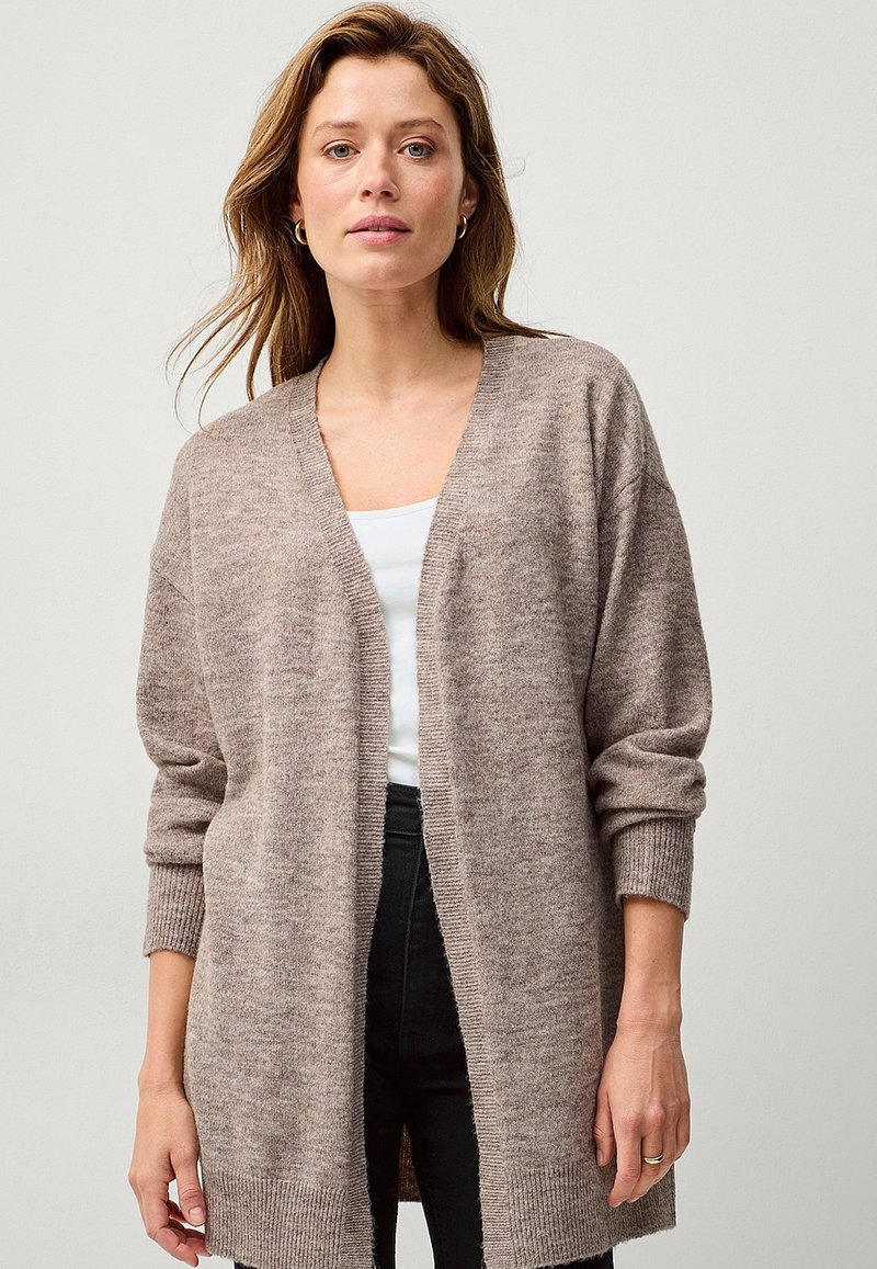 Dlouhý béžový cardigan s výstřihem do V a vzorovanými manžetami, vyrobený z měkkého strukturovaného materiálu, s otevřeným předním dílem a střední délkou, nošený přes bílý top.