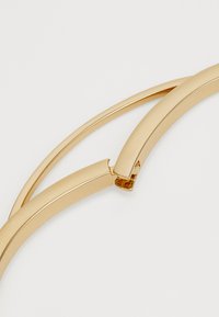 Bracciale in metallo color oro con un design liscio e curvato, caratterizzato da sezioni interconnesse e una finitura lucida. Forma minimalista e slanciata.
