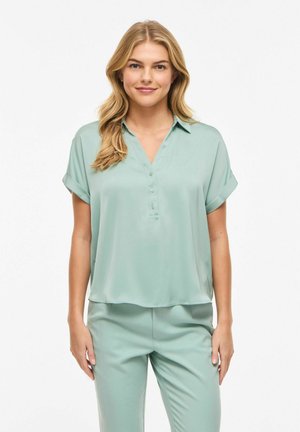 Femme blonde aux cheveux ondulés lâchés, portant un chemisier vert menthe à manches courtes boutonné et un pantalon assorti, debout devant un fond blanc.