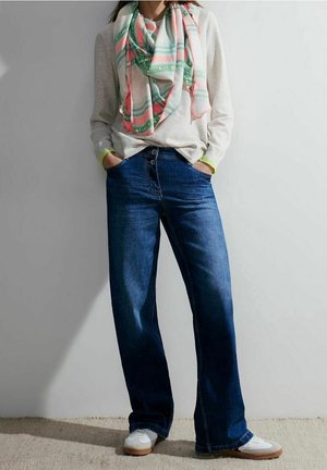 Personne portant un jean bleu à jambes larges, des baskets blanches, un pull beige clair avec un détail néon, et une écharpe à carreaux multicolore, mains dans les poches.