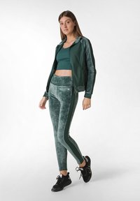 Giacca verde in velluto con zip e cappuccio, top corto abbinato e leggings a vita alta con righe verticali e dettagli testurizzati. Sneakers nere.