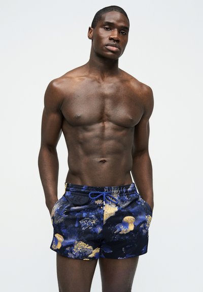 Μυώδης άνδρας με σκουρόχρωμο δέρμα που φοράει σκούρα μπλε μαγιό με χρυσά και μπλε σχέδια κοραλλιών και μέδουσες, στέκεται με τα χέρια στις τσέπες.