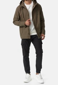 Chaqueta capucha de color verde oliva con cordones, cierre de cremallera y dos bolsillos laterales. Combinada con pantalones jogger negros y zapatillas blancas.