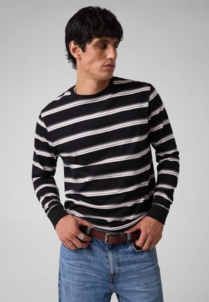 Mann trägt ein schwarz-weiß gestreiftes Langarmshirt, blaue Jeans, braunen Gürtel und schaut zur Seite, seine Hände sind nahe der Gürtelschnalle.