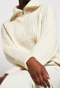 Krémově zbarvený žebrovaný pletený svetr s vysokým límcem a zapínáním na půlku zipu, s dlouhými rukávy a volným střihem. Měkká textura.