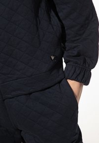 Gequiltte zwarte stoffen outfit met elastische manchetten en een zijzak, gedetailleerd met een klein hartvormig accent.