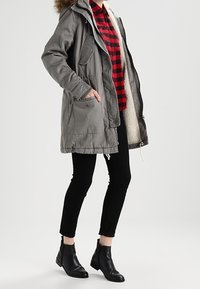 Grå parkas med en pälsfodrad huva, med framfickor och dragkedja, bärs över en röd och svart rutiga skjorta och svarta byxor.