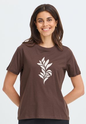 Femme aux cheveux foncés portant des boucles d'oreilles créoles dorées et un t-shirt marron avec un motif abstrait de feuille blanche, souriante, les mains dans le dos.