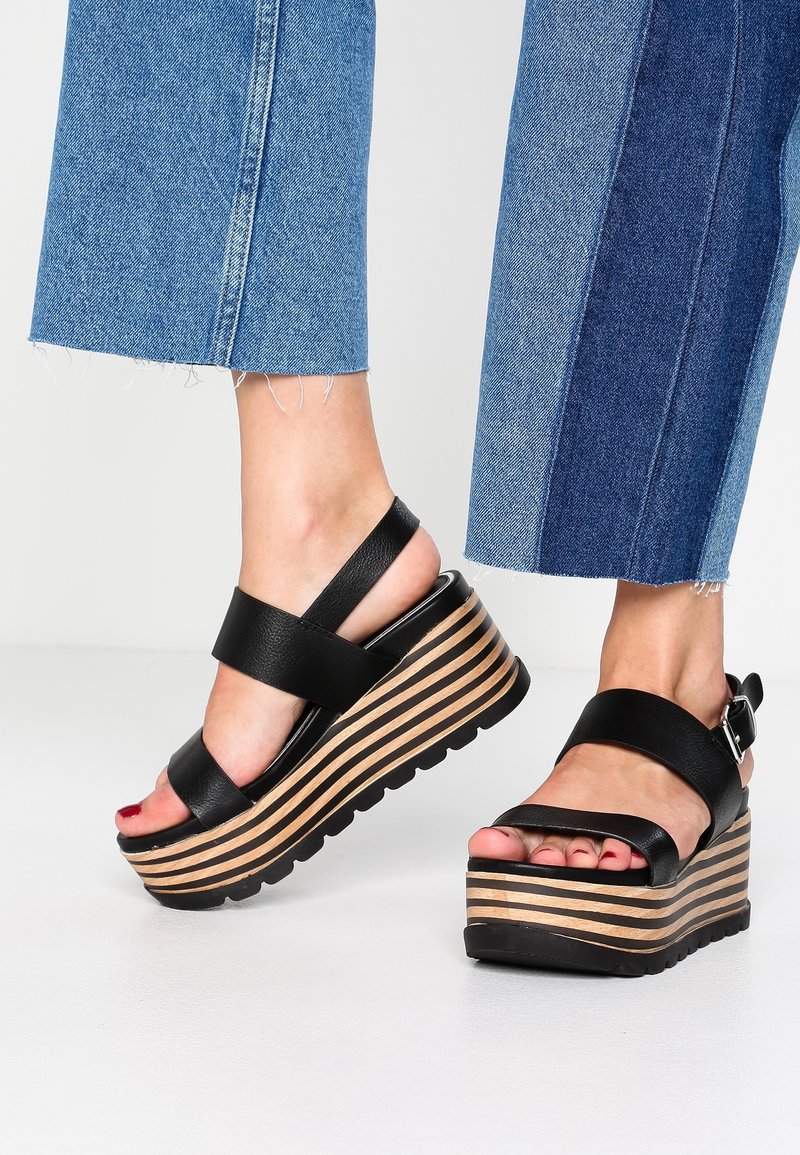 Pieds portant des sandales plateformes en cuir noir avec semelles en bois rayées, associées à un jean en denim bleu bicolore coupé court.