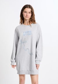 Donna che indossa un vestito felpa oversize grigio chiaro con la scritta "AMO IL MIO GATTO" e un'immagine di un gatto con un fiocco blu sulla parte anteriore.