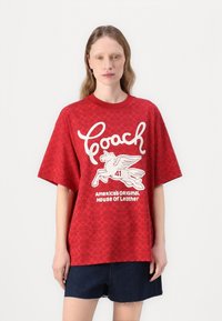 T-shirt vermelha com um corte oversized, mangas curtas e padrão texturizado; impressão gráfica branca com um cavalo alado e o nome da marca.