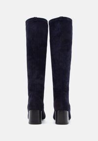 Térdmagasságú navy kék velúr csizmák sima textúrával, tömbsarkúval és minimális dizájn elemekkel a szárán.