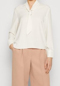 Bílá blůza s dlouhým rukávem a detailem na vázání u krku, vyrobená z hladké látky. Slučuje se s vysokými, volnějšími kalhotami v jemně růžové barvě.