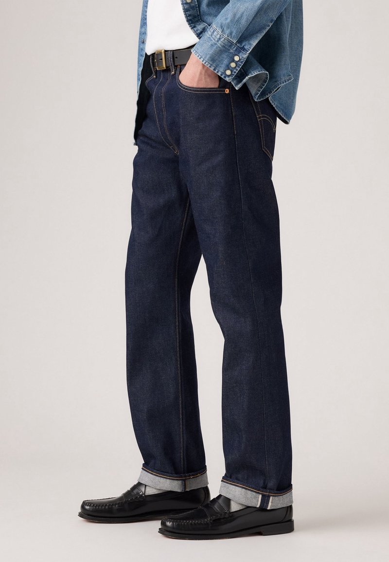 Personne portant un jean droit bleu foncé avec ourlets retroussés, ceinture noire, veste en denim, chemise blanche, chaussettes grises et mocassins en cuir noir.
